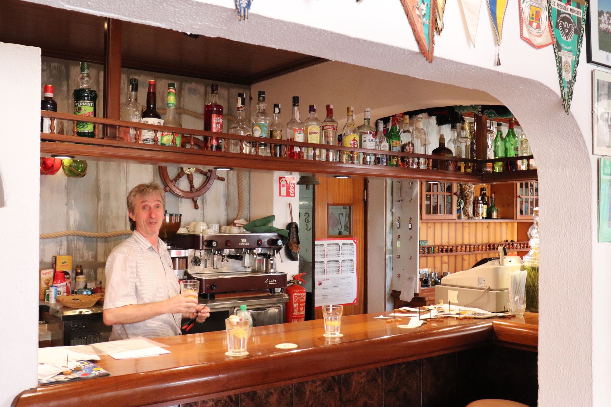 A member of the family pouring behind the wooden bar, shelves of bottles and glasses around