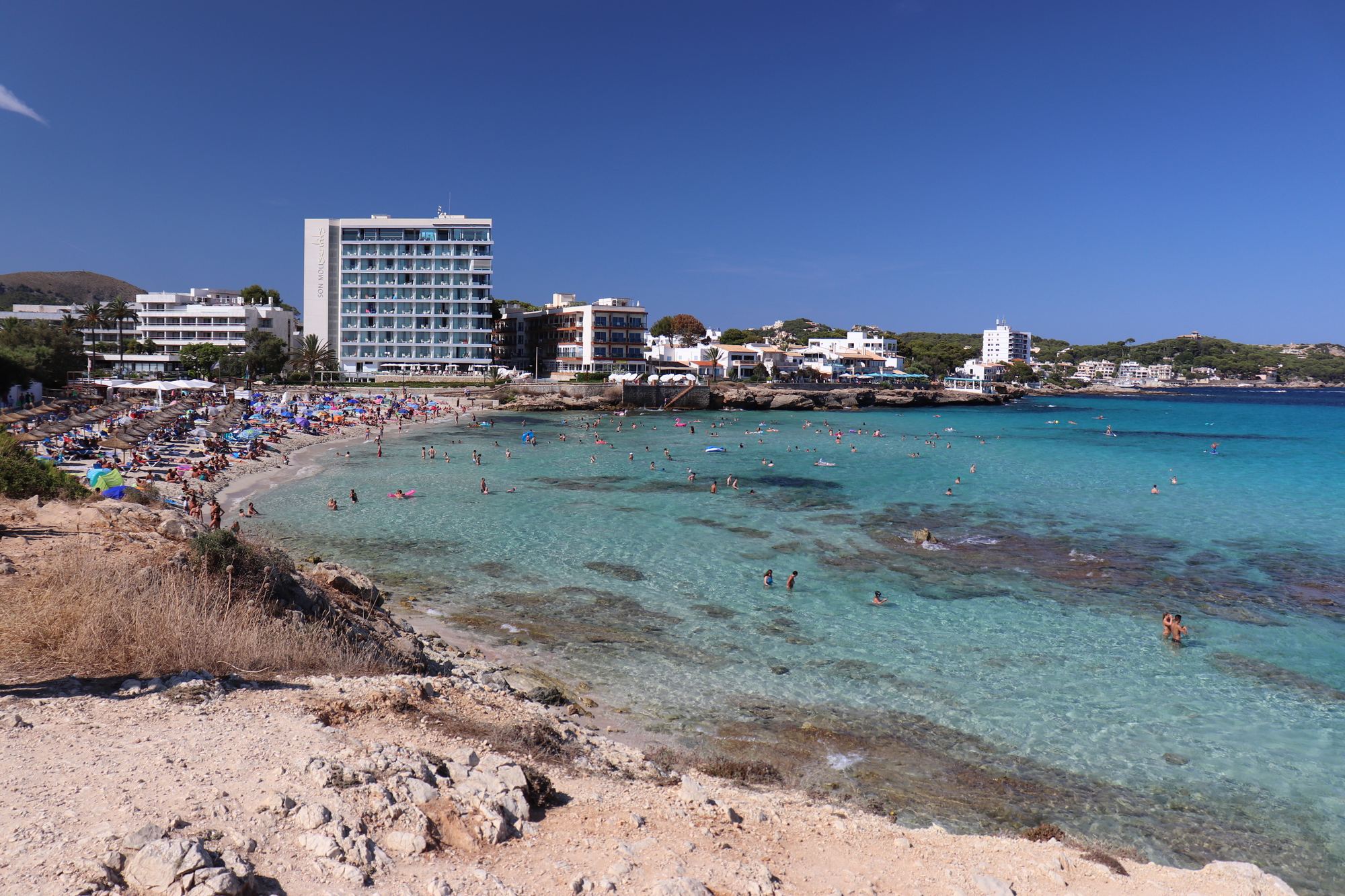 A high view over a Cala Ratjada bay: turquoise shallows fading to deep blue, a busy strip of sand and the white town along the headland