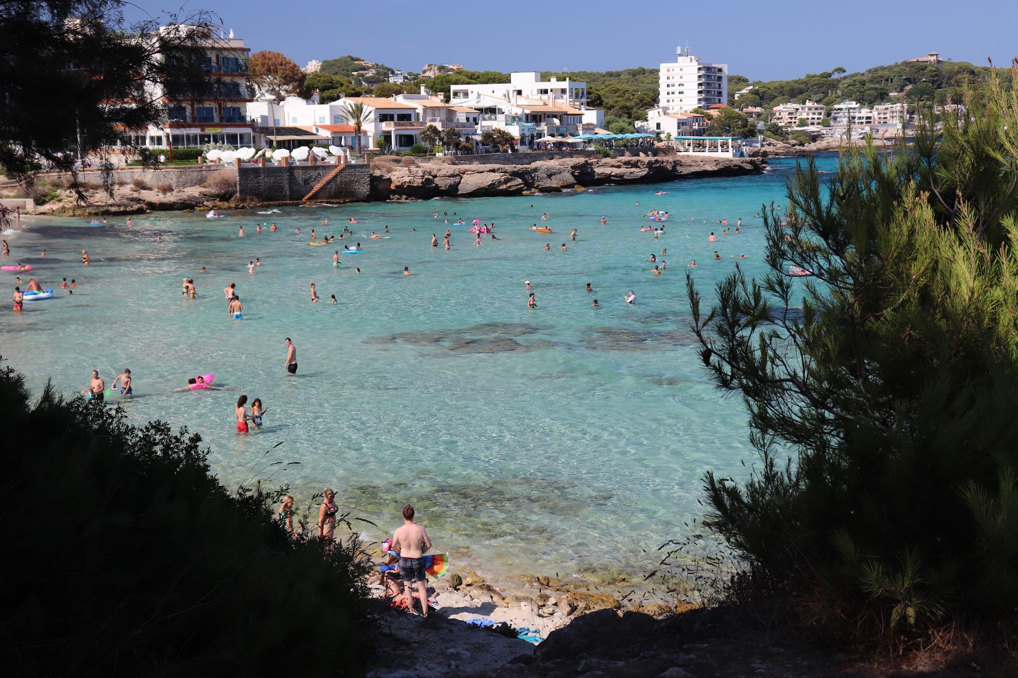 Son Moll town beach, swimmers in clear shallow water with the white buildings of Cala Ratjada along the shore