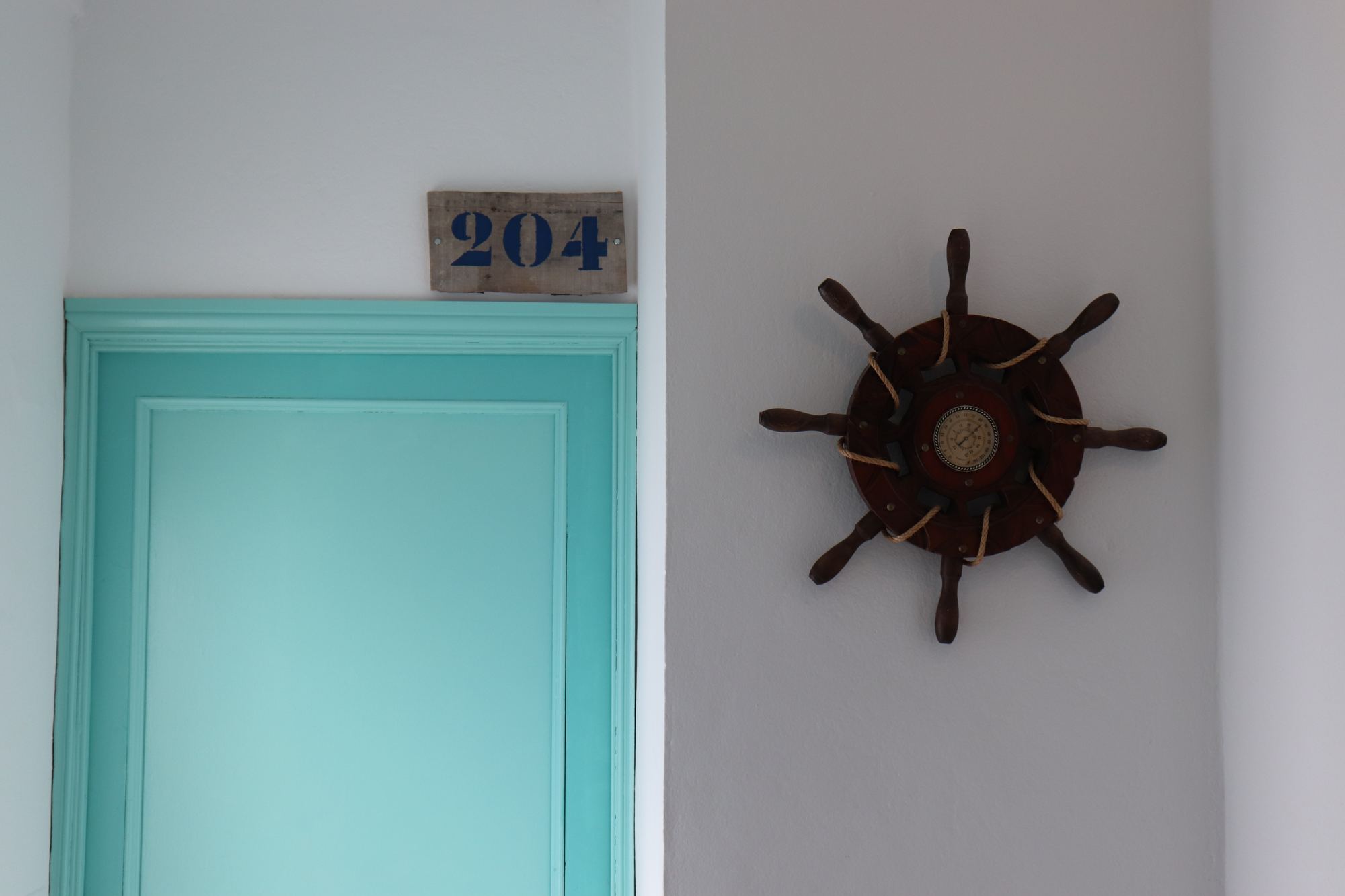 A turquoise-painted room door under a small numbered 204 plaque, with a dark wooden ship's wheel mounted on the white wall beside it