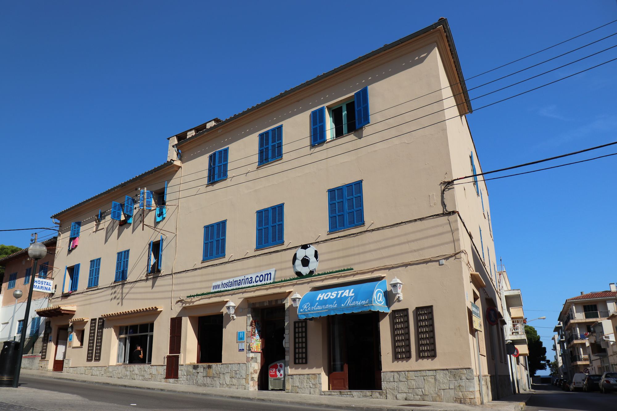 Hostal Marina seen along its street: the long sandstone front with rows of blue shutters and the blue HOSTAL MARINA sign, palms and a clear Mallorcan sky above