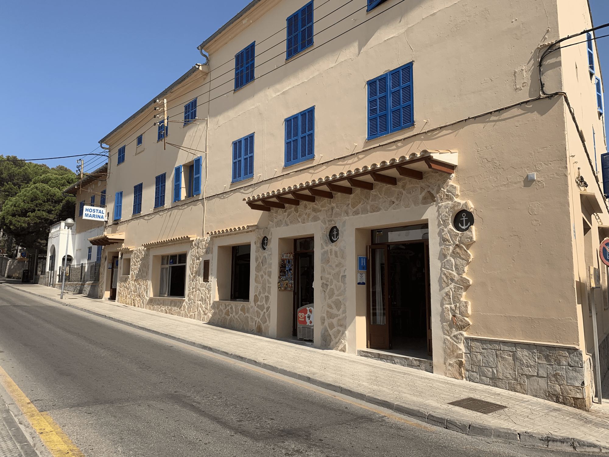 The sandstone façade of Hostal Marina on its quiet Cala Ratjada street, three storeys of cobalt-blue shutters above a stone-framed ground floor with a timber porch and a brass anchor by the door