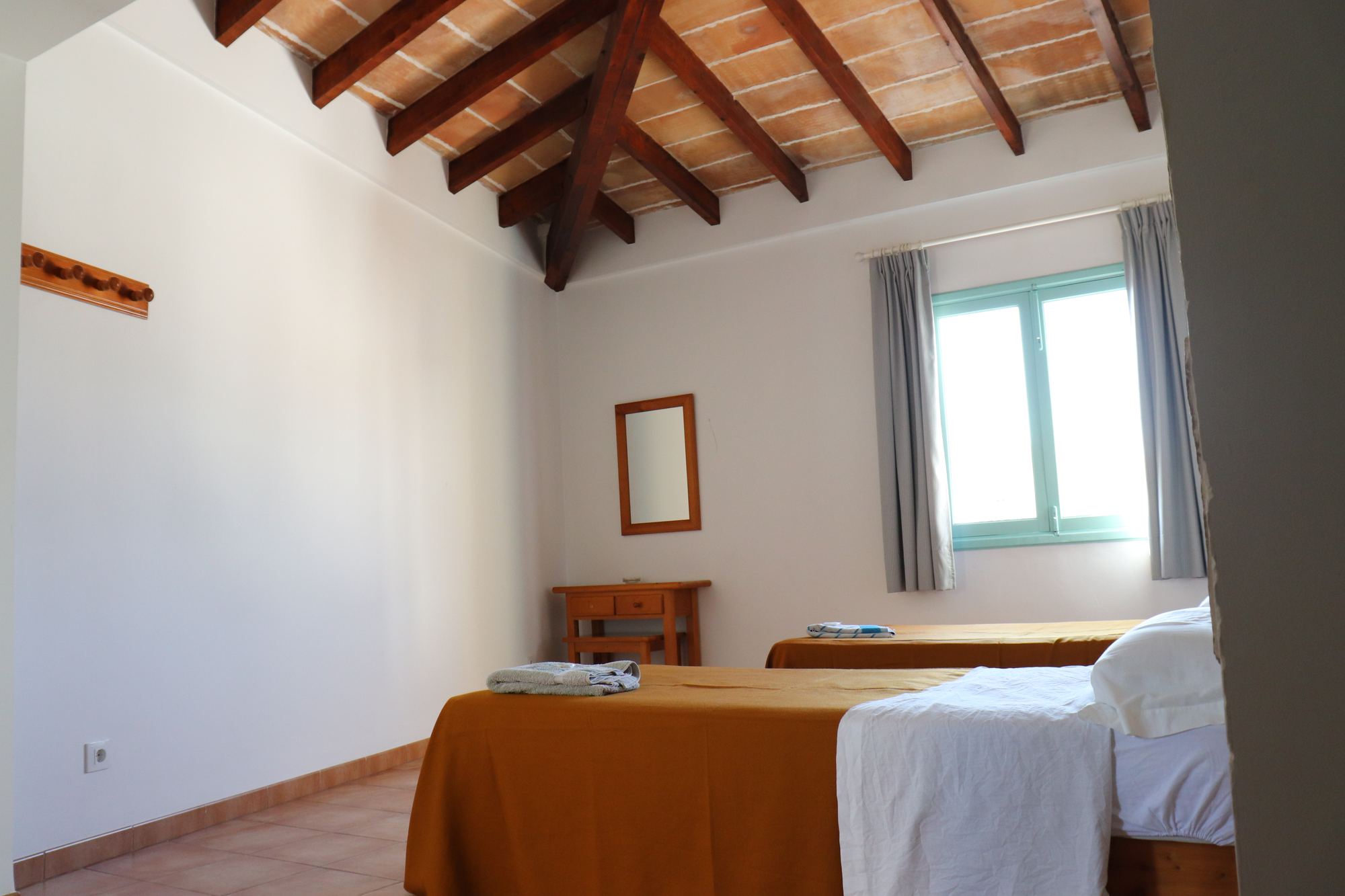 A twin room with two beds in mustard-gold covers, white walls, a wood-beamed ceiling and a small window in a blue frame