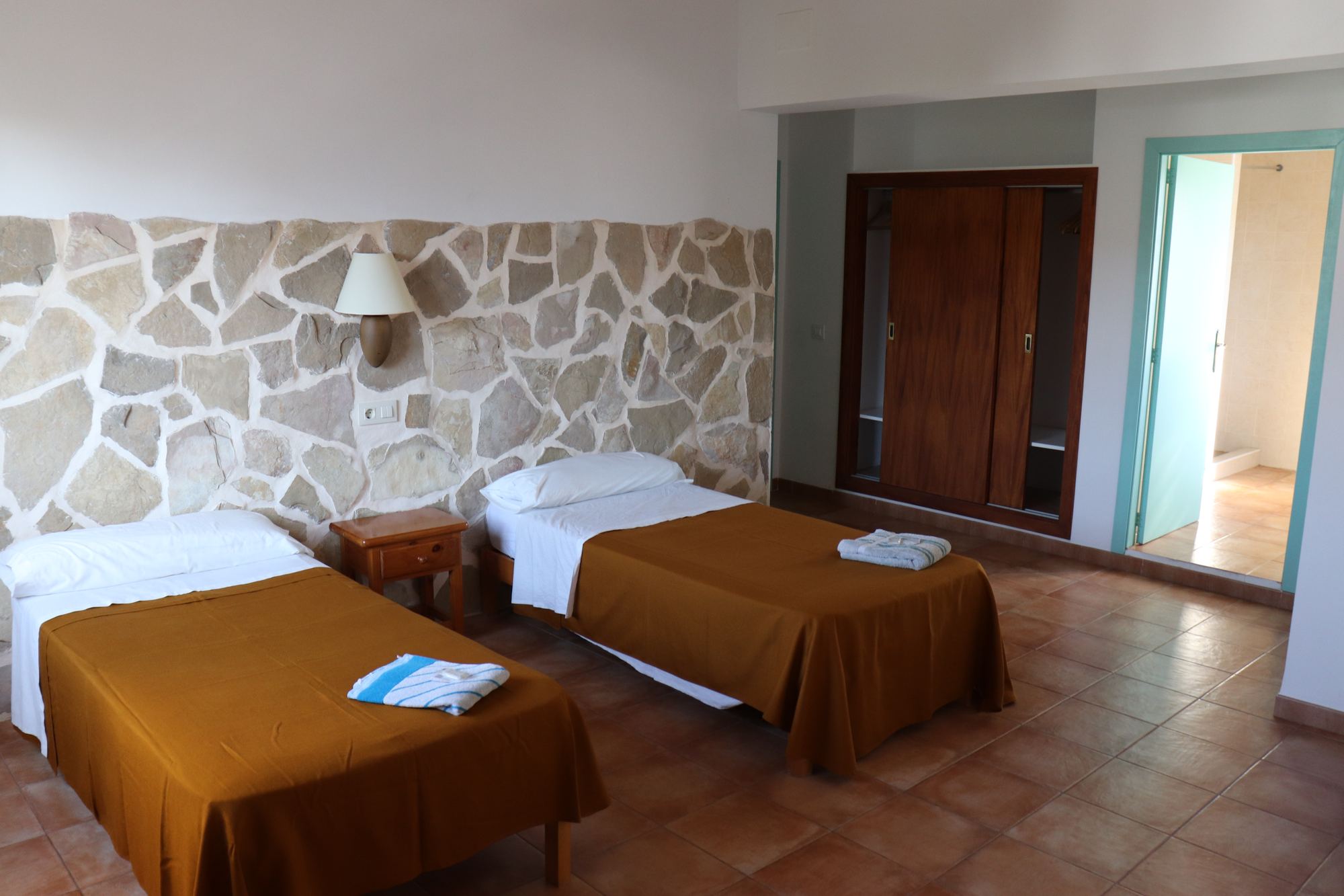 A twin room with a natural stone feature wall, two beds in gold covers, a tiled floor and a doorway through to a bright bathroom