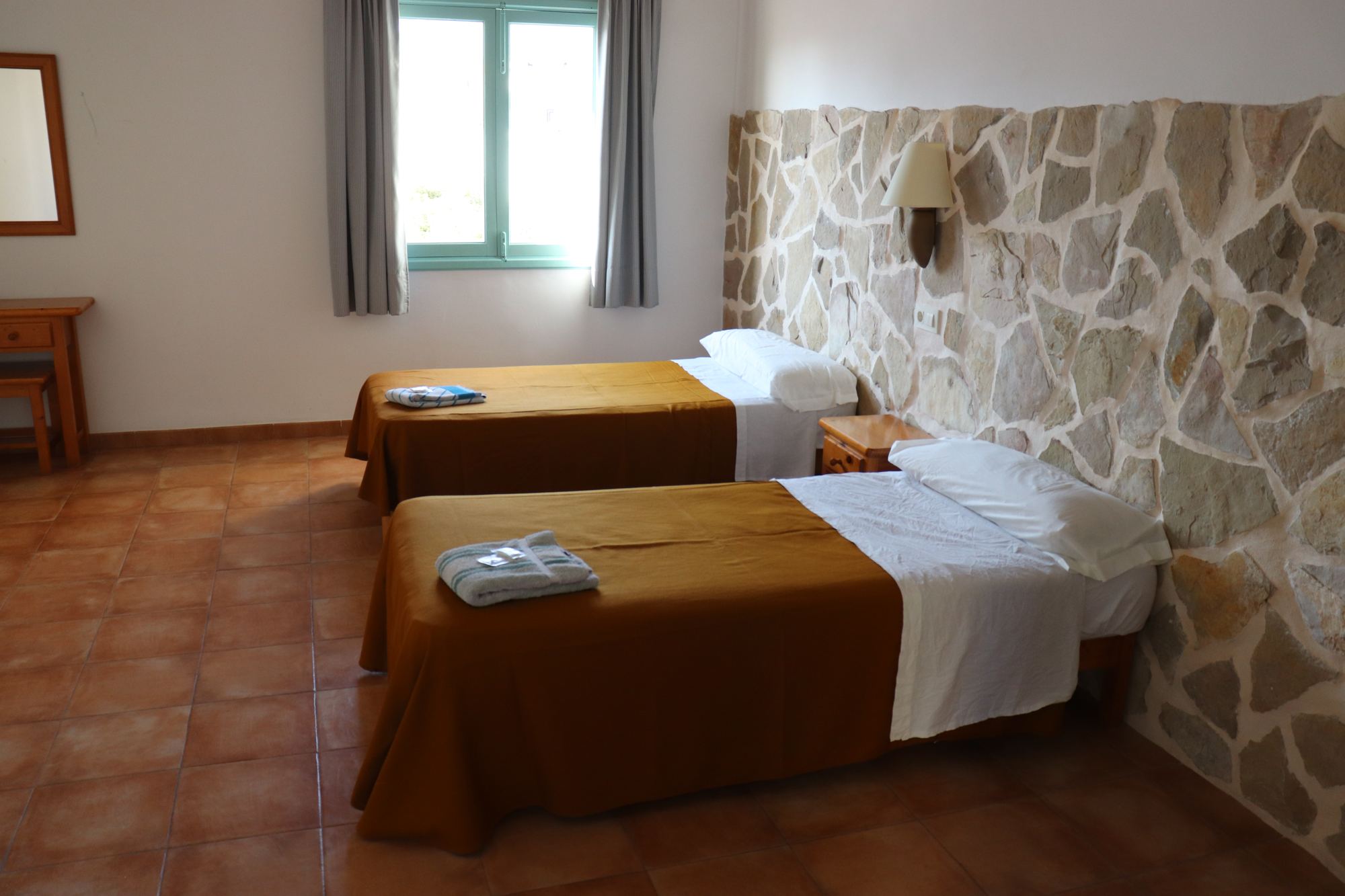 A simple twin room with white walls, two single beds in gold covers, a wooden ceiling and warm afternoon light